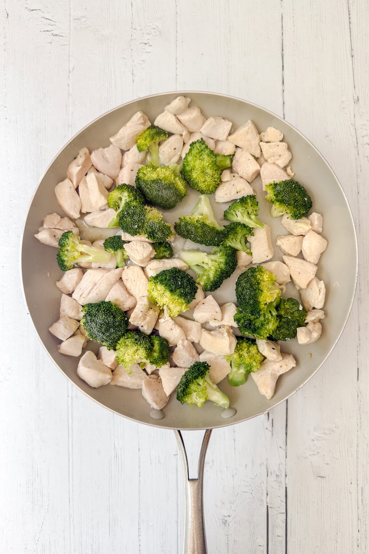 A frying pan on a white wooden surface contains delicious Chicken and Broccoli, with juicy pieces of chicken breast and fresh steamed broccoli florets evenly distributed across the pan.