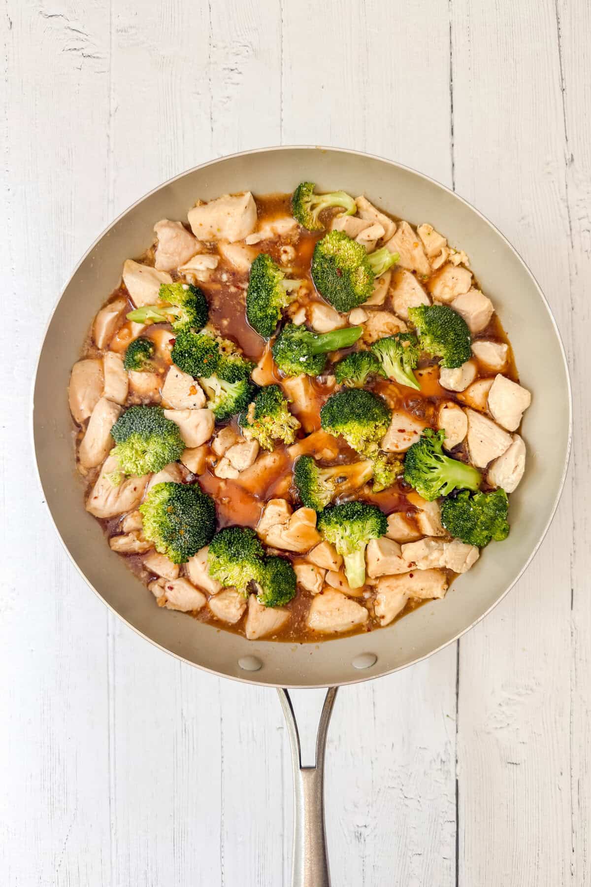 A skillet filled with tender chicken and broccoli pieces coated in a savory brown sauce, set on a white wooden surface.