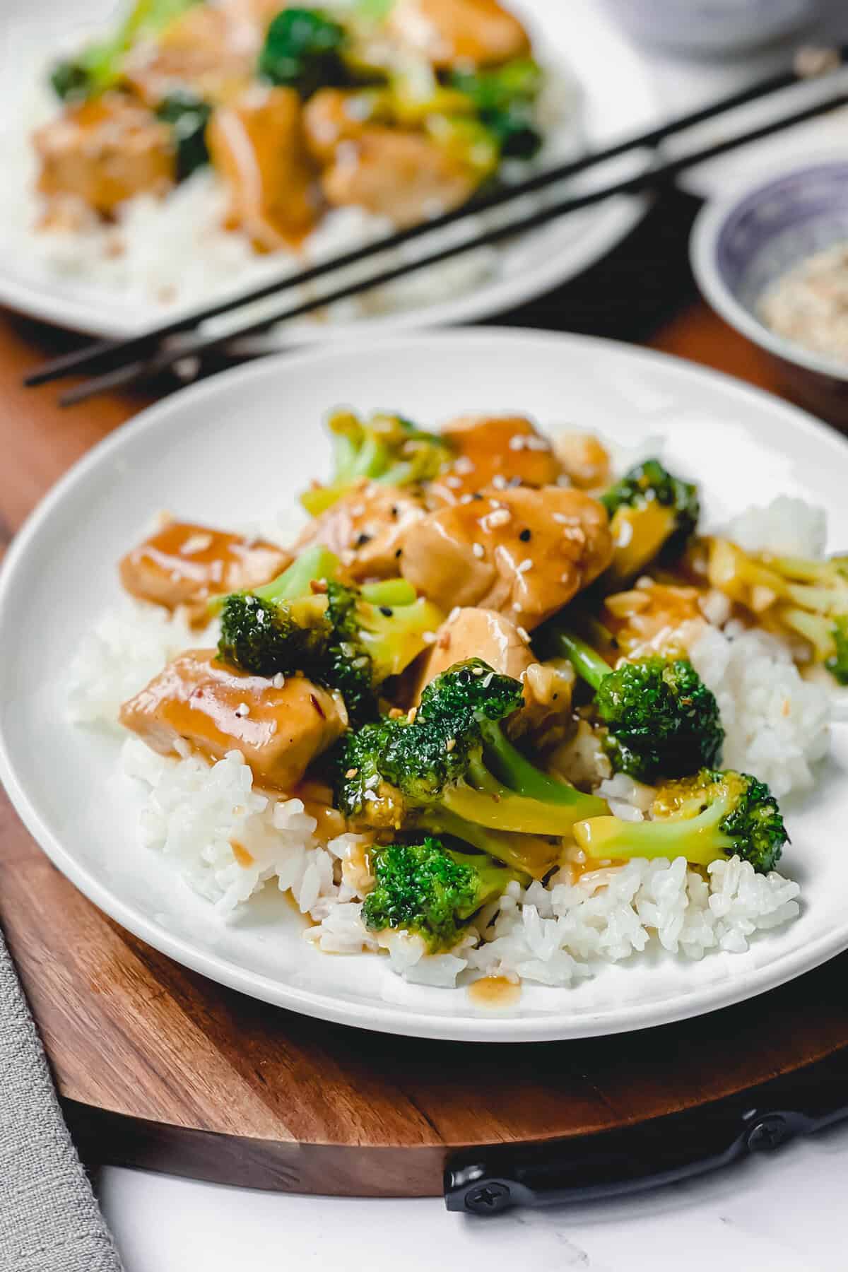 A plate of Chicken and Broccoli features tender chicken pieces and broccoli in a savory brown sauce over white rice, served on a white plate with chopsticks, and another plate of food in the background.