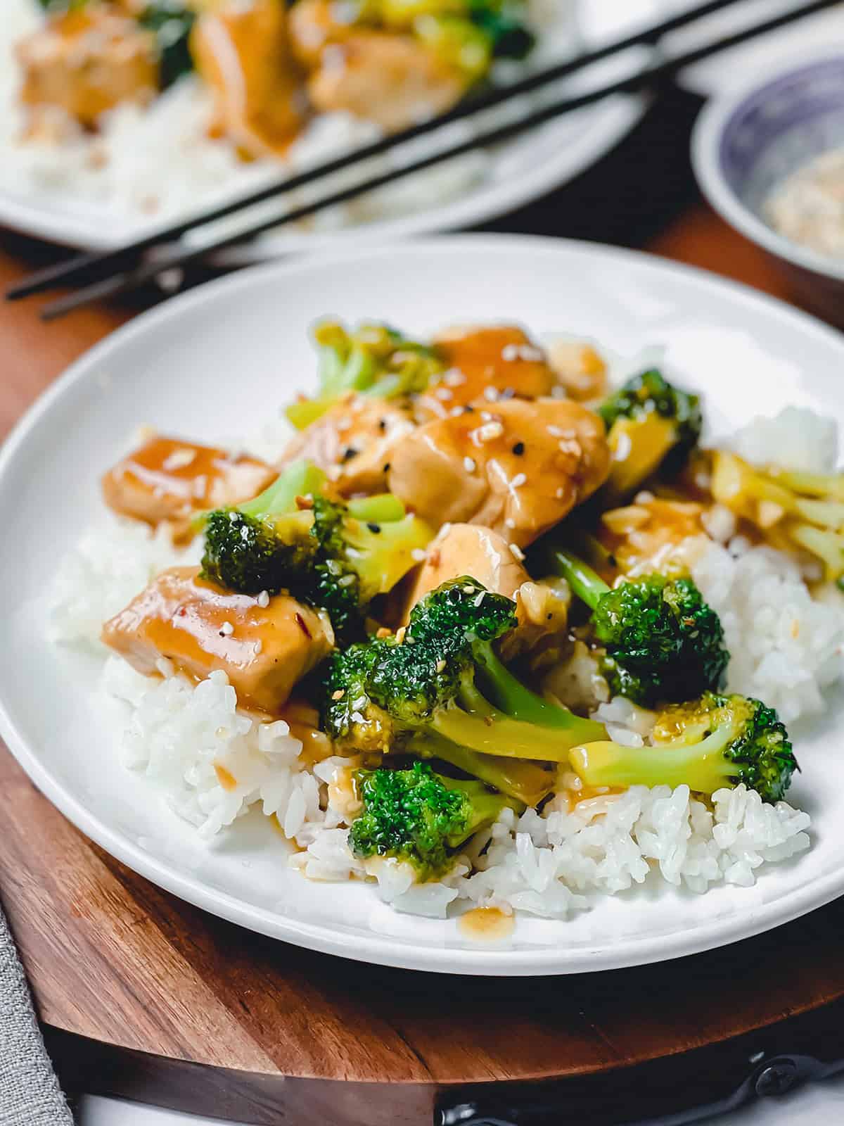 A white plate of Chicken and Broccoli served over rice, topped with savory brown sauce. Chopsticks and another plate can be seen in the background.