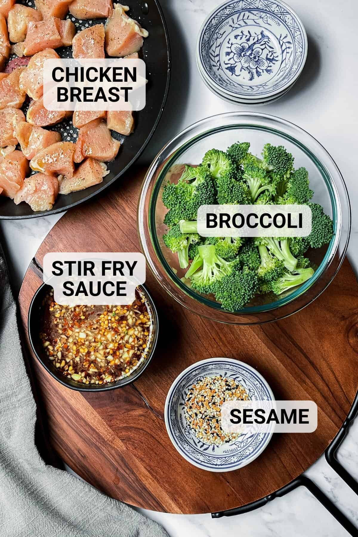 Top-down view of a wooden board holding bowls of raw chicken and broccoli florets, stir fry sauce, and a small dish of sesame seeds, each labeled with text. Perfect for making a classic Chicken and Broccoli stir fry.