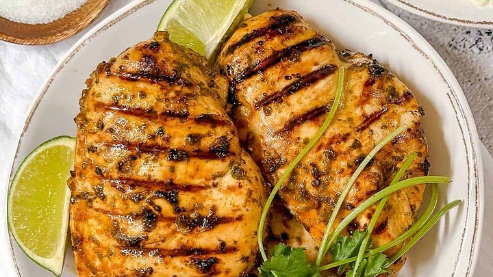 Top view of grilled Cilantro Lime Chicken breasts on a white plate with lime wedges, fresh cilantro and rice in the background.