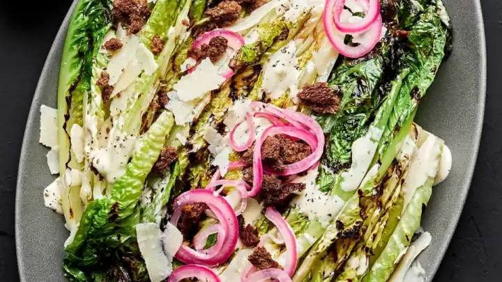 Oval platter with grilled romaine lettuce, Caesar dressing, shaved parmesan, pickled red onions, and crumbled dark topping—perfect for fans of grill recipes—surrounded by small bowls of toppings and extra dressing.