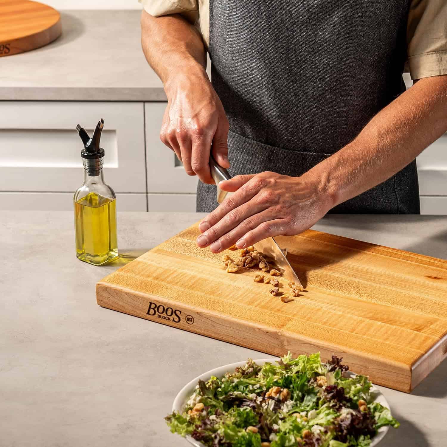 A person chops nuts on a wooden cutting board next to a bottle of olive oil and a bowl of salad, preparing delicious ingredients for fresh salad recipes on the kitchen counter.