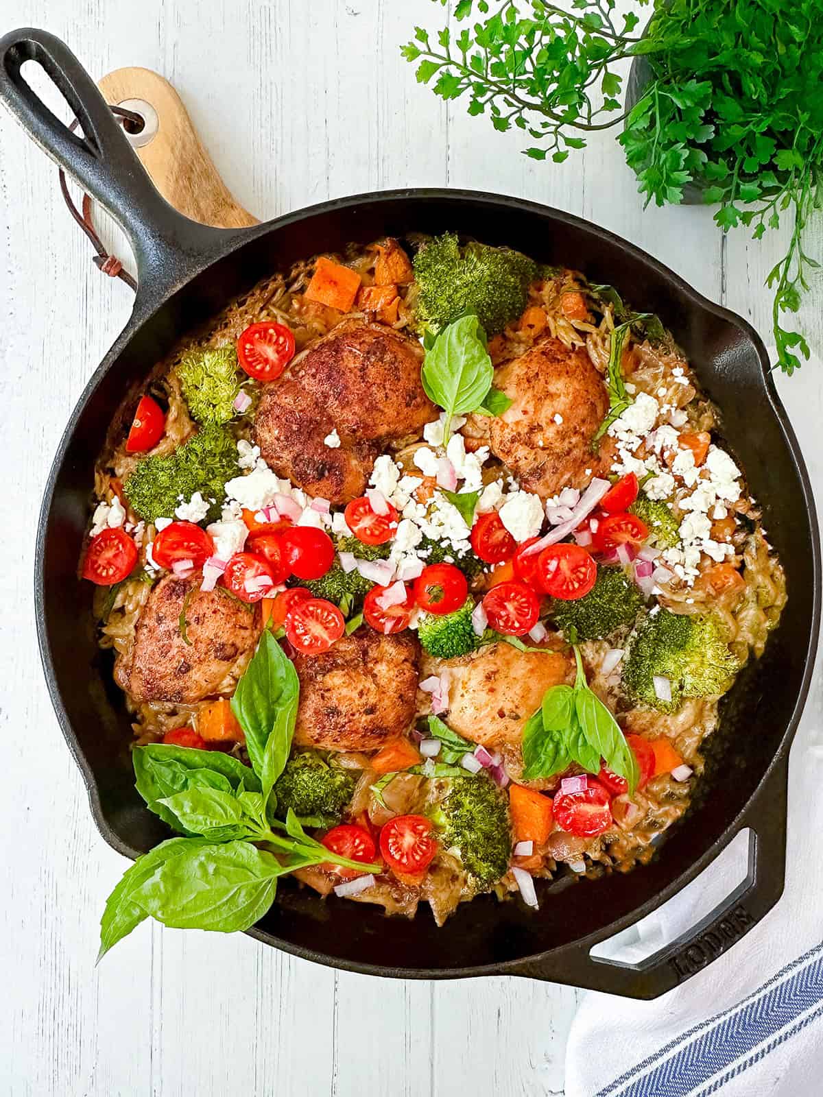 A cast iron skillet filled with Mediterranean chicken, broccoli, sweet potatoes, cherry tomatoes, feta cheese, red onion, and fresh basil sits on a white wooden surface.