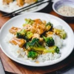 A plate of Chicken and Broccoli features white rice topped with tender chicken, crisp broccoli florets, and savory brown sauce, served on a white dish with chopsticks nearby.