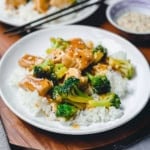 A white plate with white rice topped with tender Chicken and Broccoli pieces in savory brown sauce, with chopsticks and a small bowl in the background.