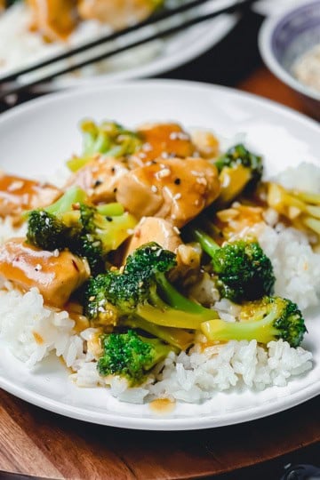 A white plate with white rice topped with tender Chicken and Broccoli pieces in savory brown sauce, with chopsticks and a small bowl in the background.