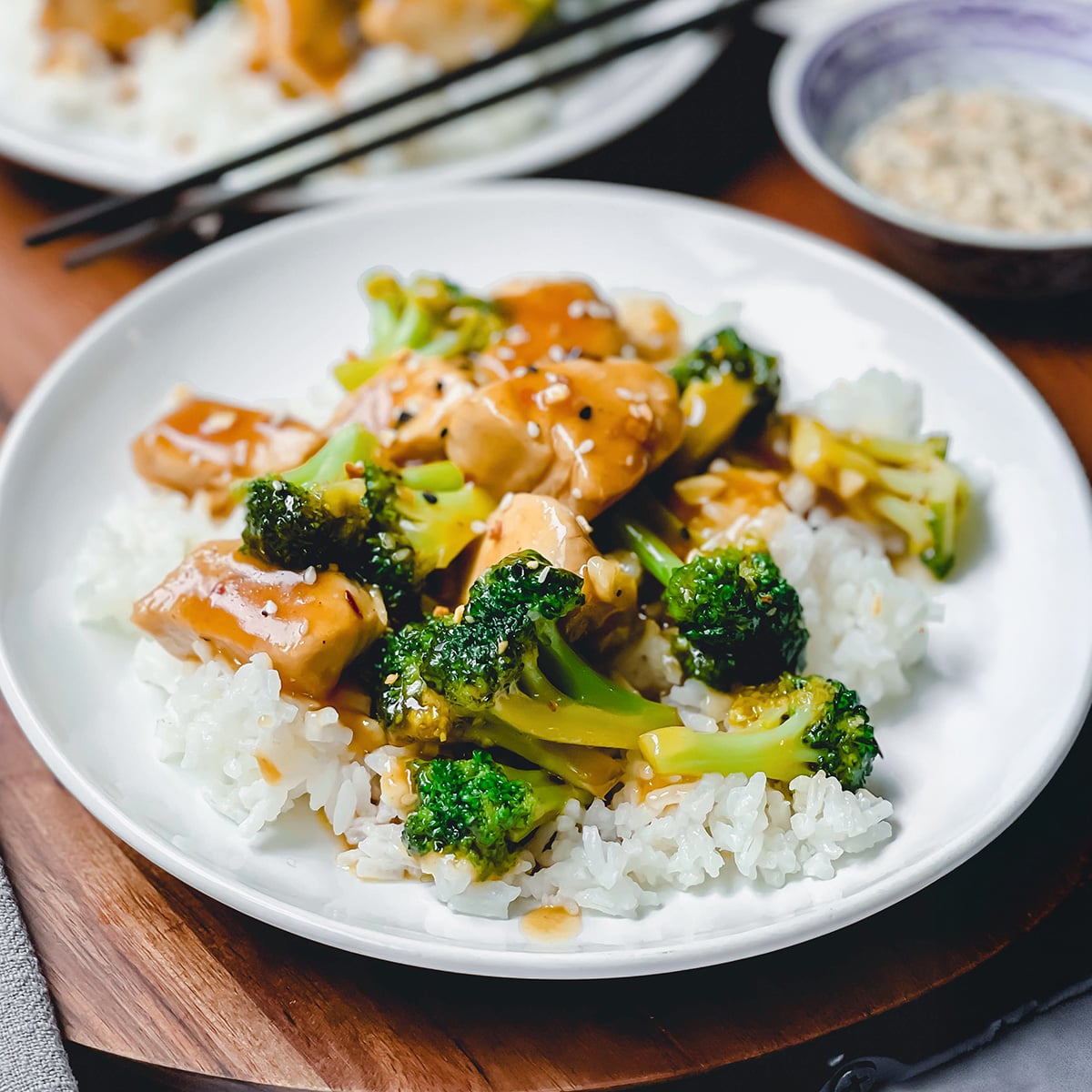 A white plate with white rice topped with tender Chicken and Broccoli pieces in savory brown sauce, with chopsticks and a small bowl in the background.