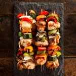 Four teriyaki chicken skewers with grilled vegetables are arranged on a rectangular slate serving platter, placed on a rustic wooden surface.