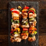 Four teriyaki chicken skewers with grilled vegetables are arranged on a rectangular slate serving platter, placed on a rustic wooden surface.