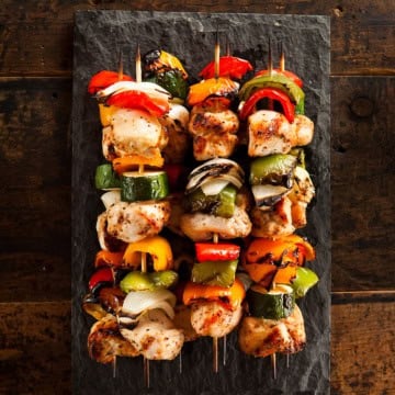 Four teriyaki chicken skewers with grilled vegetables are arranged on a rectangular slate serving platter, placed on a rustic wooden surface.