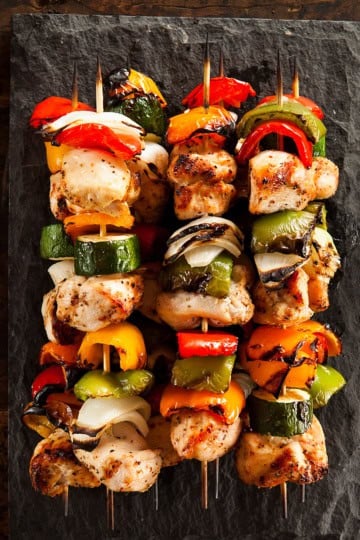 Four teriyaki chicken skewers with grilled vegetables are arranged on a rectangular slate serving platter, placed on a rustic wooden surface.