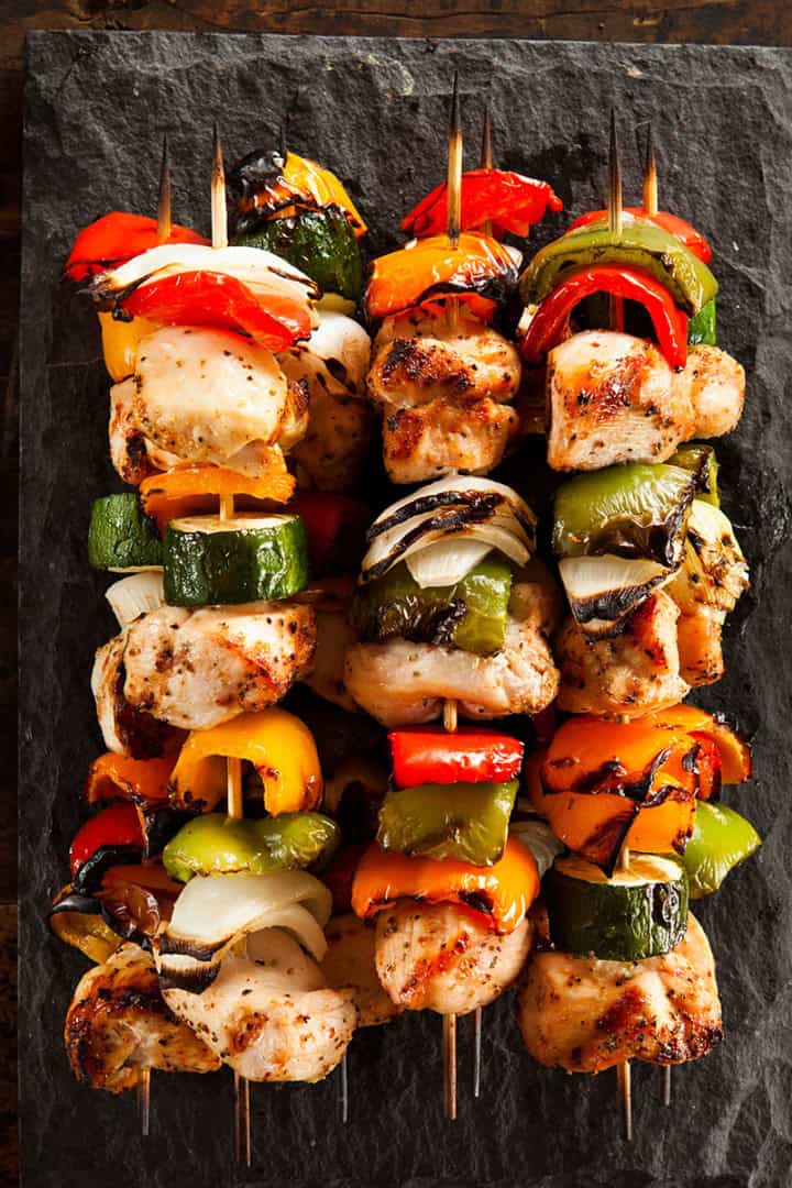 Four teriyaki chicken skewers with grilled vegetables are arranged on a rectangular slate serving platter, placed on a rustic wooden surface.