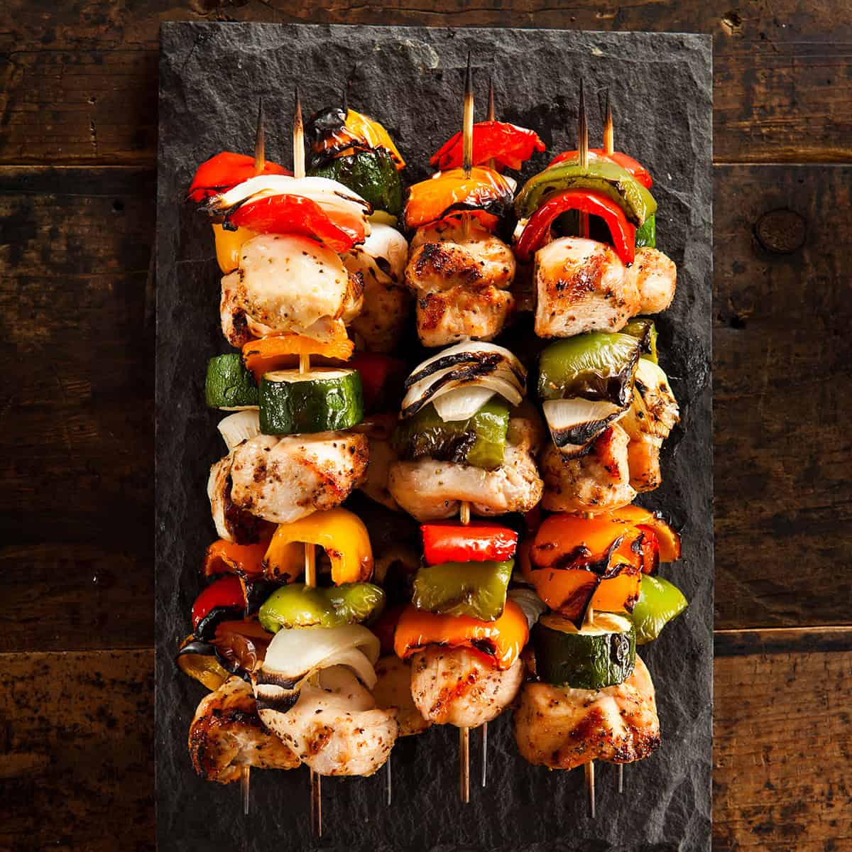 Four teriyaki chicken skewers with grilled vegetables are arranged on a rectangular slate serving platter, placed on a rustic wooden surface.