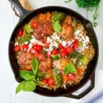 A skillet with Mediterranean chicken thighs, broccoli, cherry tomatoes, diced carrots, red onion, feta cheese, and fresh basil served on a rice base.
