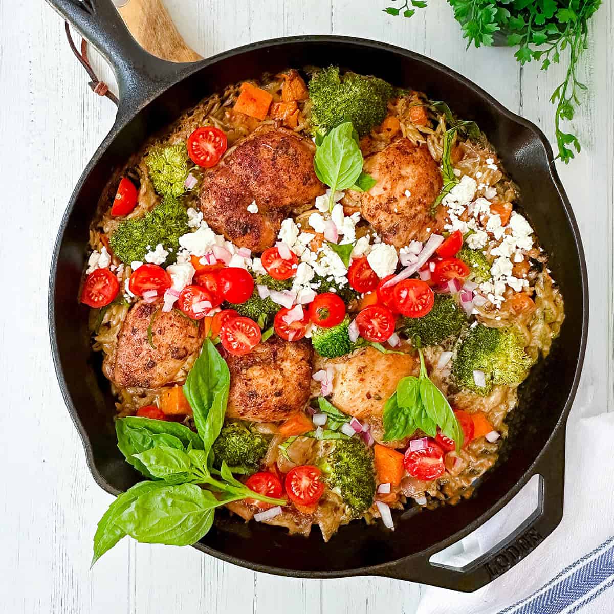 A skillet with Mediterranean chicken thighs, broccoli, cherry tomatoes, diced carrots, red onion, feta cheese, and fresh basil served on a rice base.