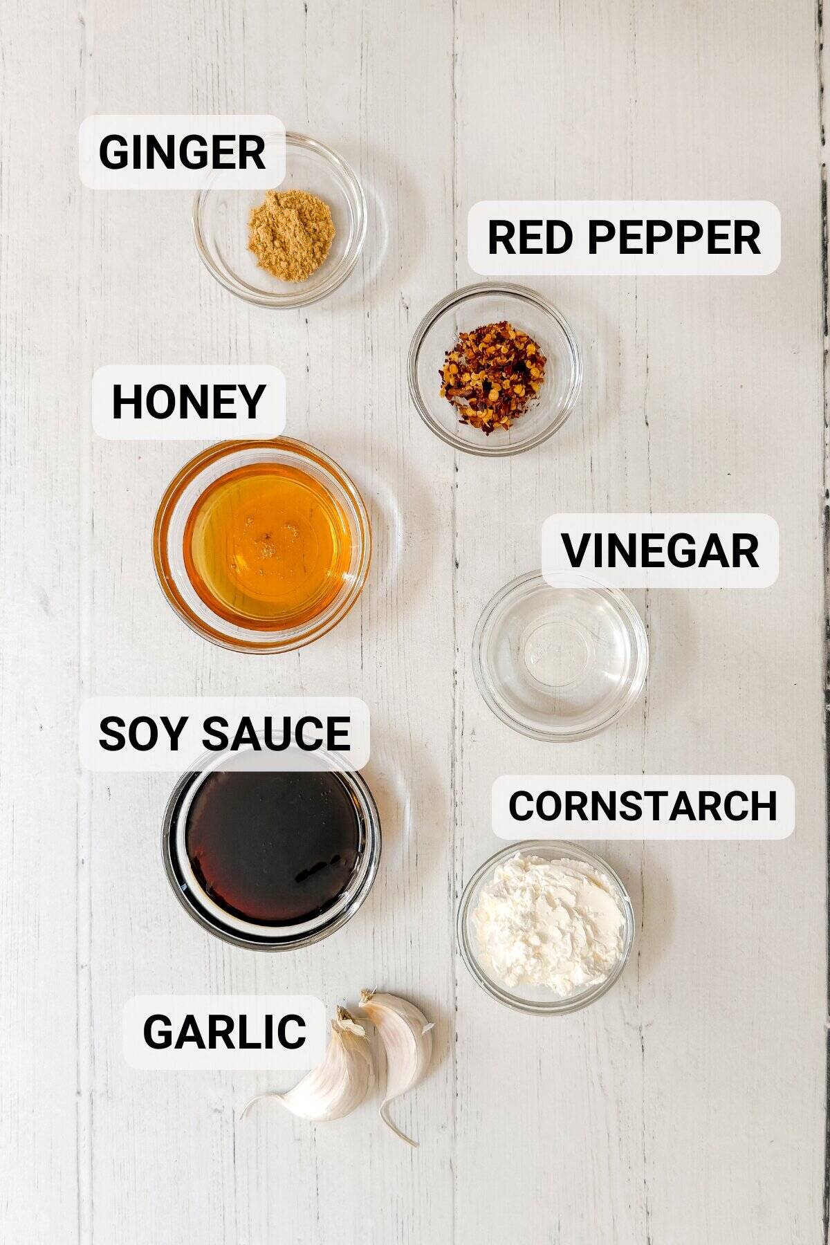 Eight labeled ingredients for a Chicken and Broccoli recipe are arranged on a white surface: ginger, red pepper, honey, vinegar, soy sauce, cornstarch, and two garlic cloves. Each ingredient is in a small bowl except the garlic.