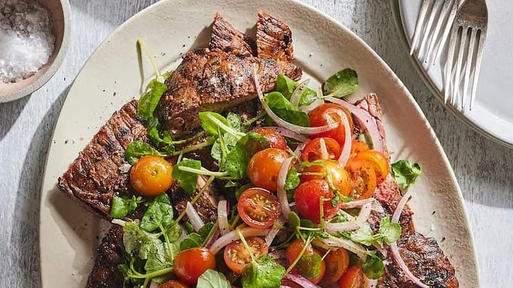 Grilled steak slices topped with a salad of cherry tomatoes, red onions, and greens on a white oval plate—a mouthwatering example of flavorful grill recipes—with salt and empty dishes nearby.