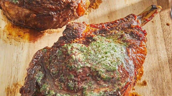 Two cooked bone-in ribeye steaks with a green herb butter topping rest on a wooden cutting board, making them a delicious highlight for your favorite grill recipes.