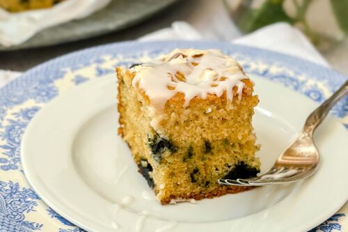 Square of iced Almond Blueberry Cake on a white plate with a fork.