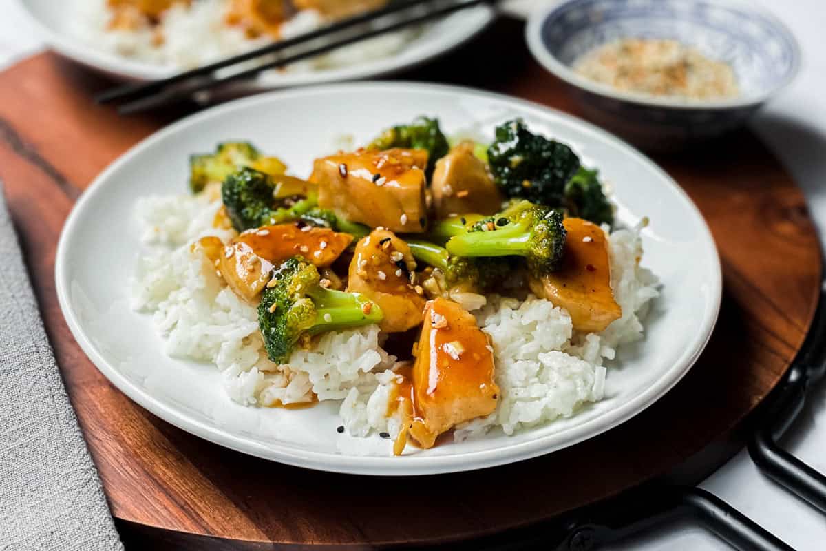 A plate of Chicken and Broccoli features tender chicken, crisp broccoli, and glossy brown sauce over white rice, garnished with sesame seeds. A bowl with seasoning and chopsticks are in the background.