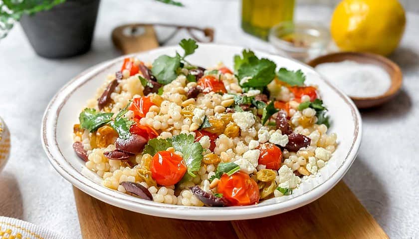 Horizontal side view of Israel Couscous (Pearl Couscous) in a white serving bowl with fresh herbs and toasted pine nuts.