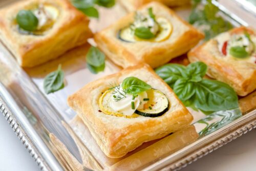 Horizontal closeup view of puff pastry appetizer served on a silver tray with fresh vegetables and herbs.