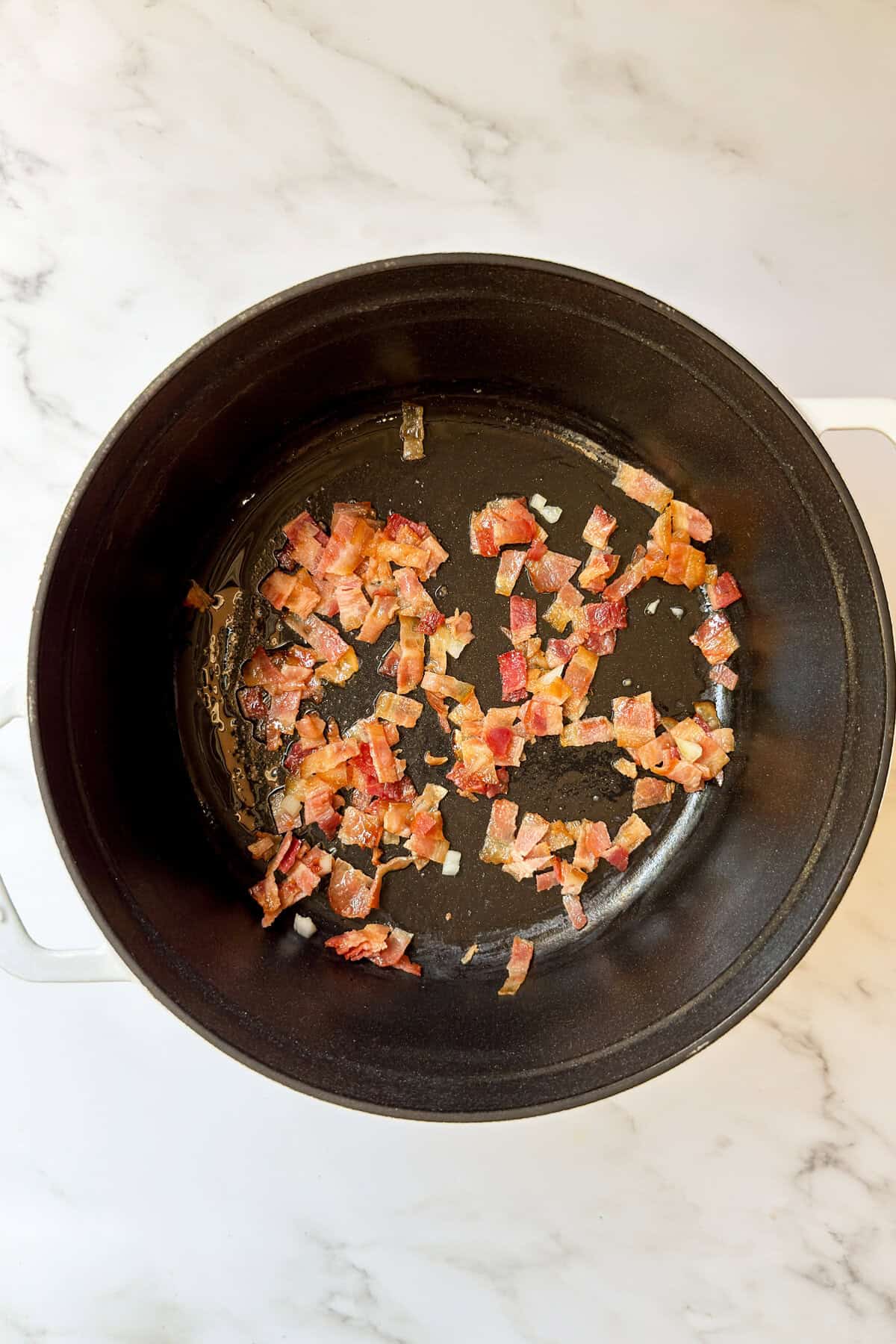 Chopped bacon pieces are being cooked in a black pot on a white marble countertop as the first step in preparing a hearty corn chowder recipe.