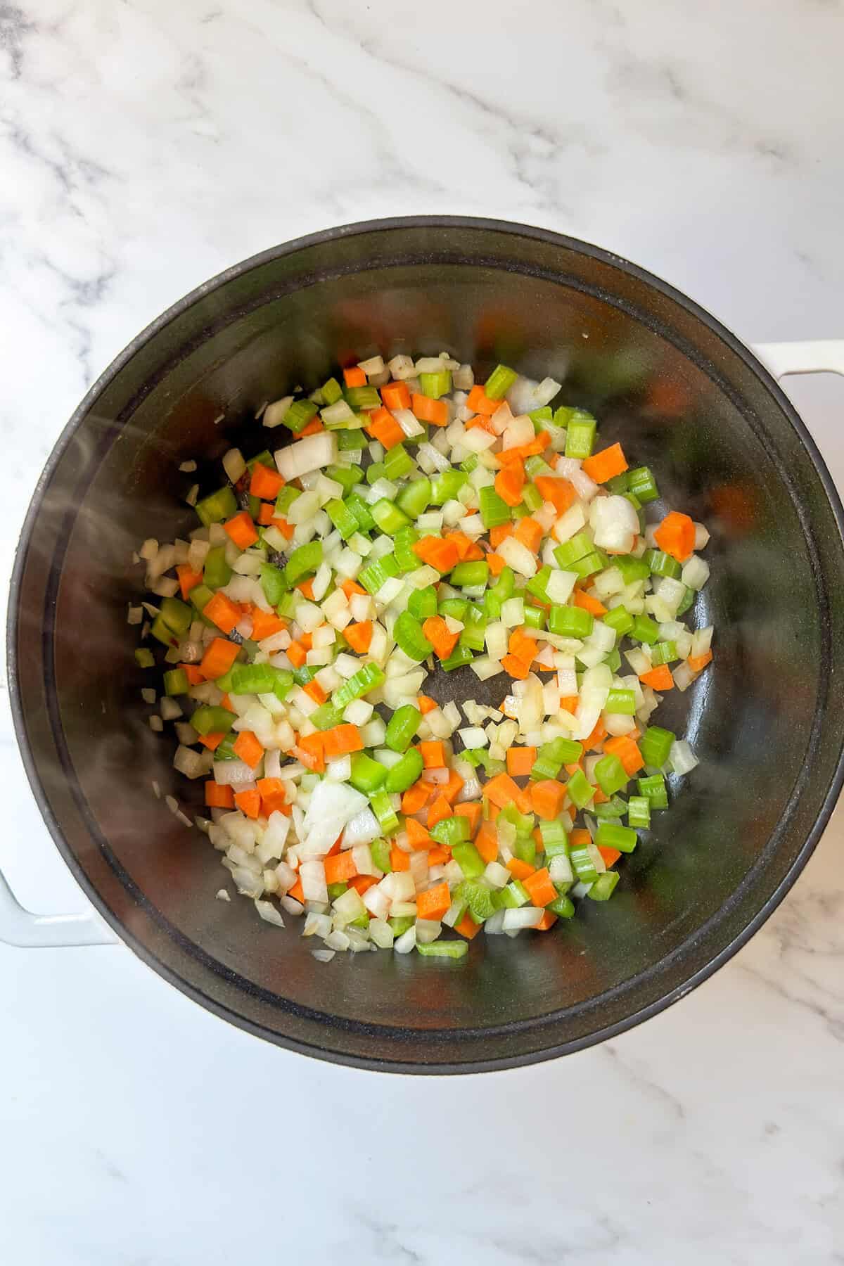 A pot filled with chopped onions, carrots, and celery sautéing on a white marble countertop-the perfect base for a comforting corn chowder recipe.
