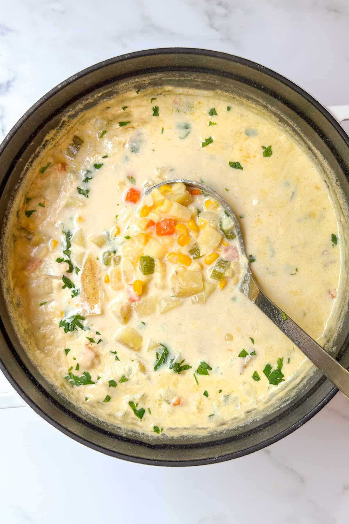A pot of creamy corn chowder recipe with a ladle, filled with diced potatoes, corn, carrots, and herbs, sits on a white marble surface. The soup is garnished with chopped parsley.