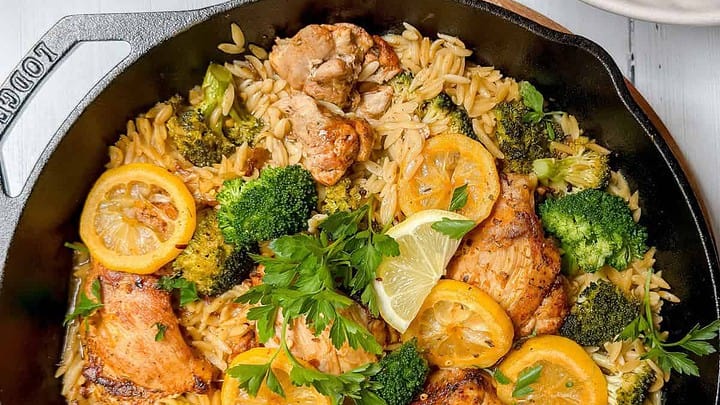 A skillet filled with cooked orzo, broccoli, lemon slices, and pieces of chicken, garnished with fresh parsley and lemon wedges—perfect for August Mediterranean Diet Dinners.