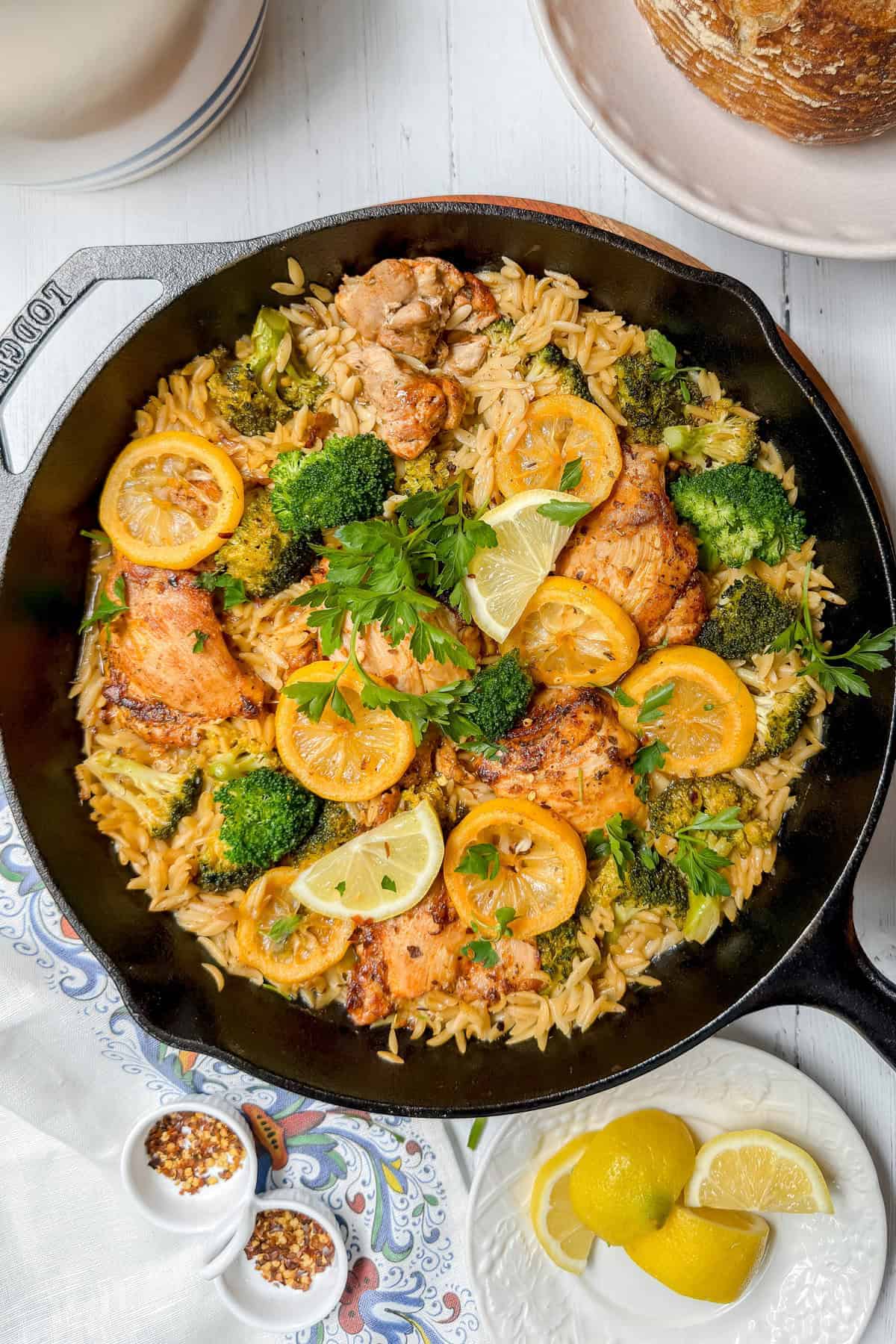 A skillet filled with cooked orzo, broccoli, lemon slices, and pieces of chicken, garnished with fresh parsley and lemon wedges-perfect for August Mediterranean Diet Dinners.