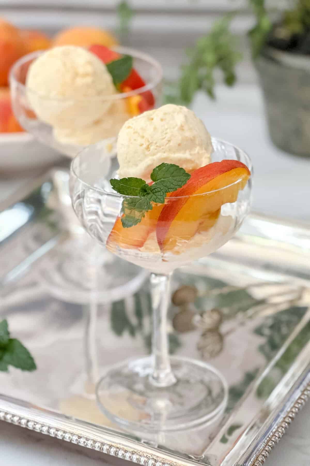 Closeup view of two scoops of peach ice cream in a dessert goblet with fresh peach slices and mint sprigs.