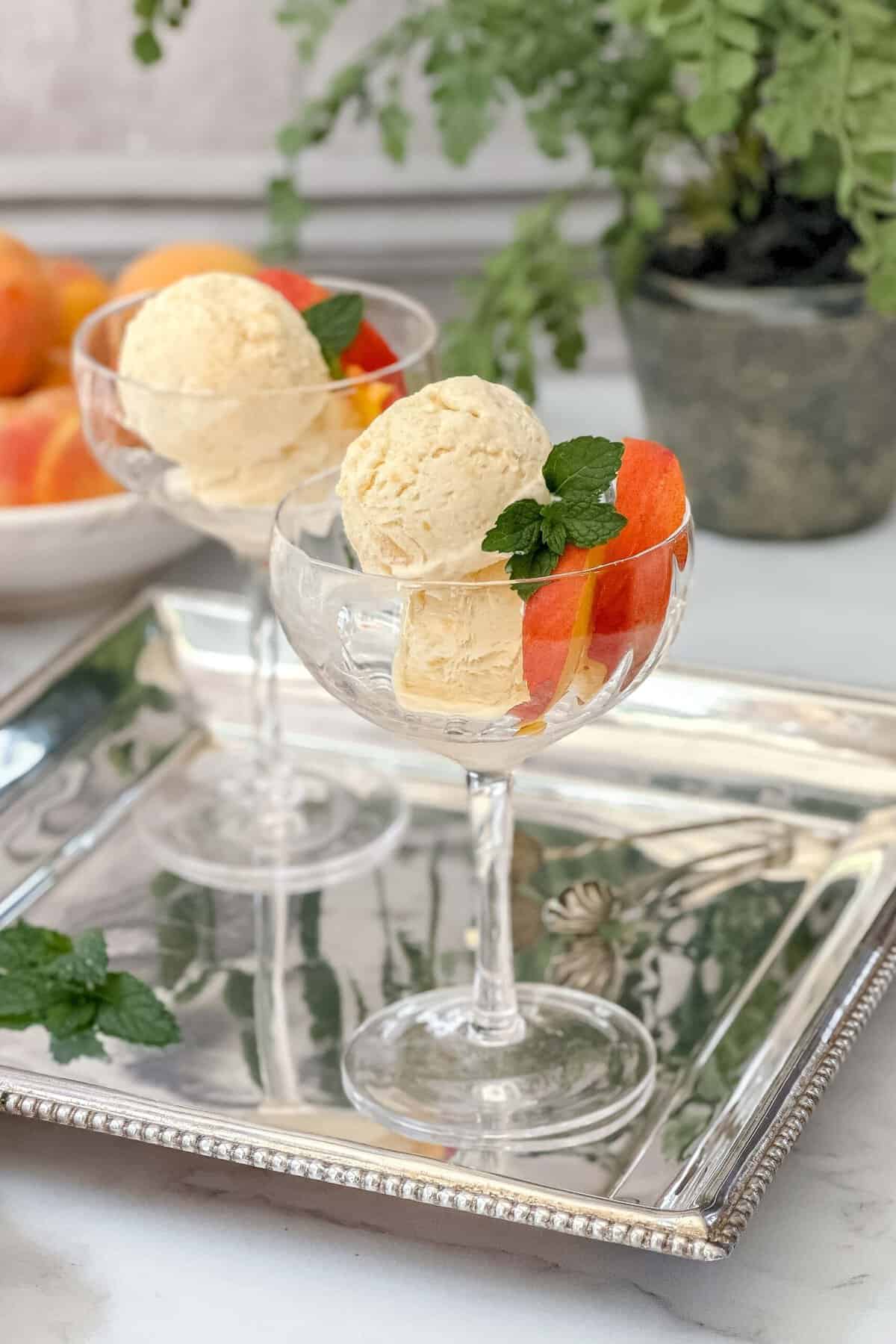 Side vertical view of peach ice cream in a tall dessert goblet with fresh peaches and mint sprigs.