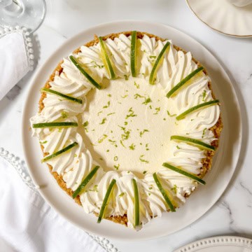 A ricotta cheesecake inspired key lime pie topped with whipped cream, lime wedges, and lime zest sits on a white plate on a marble surface.