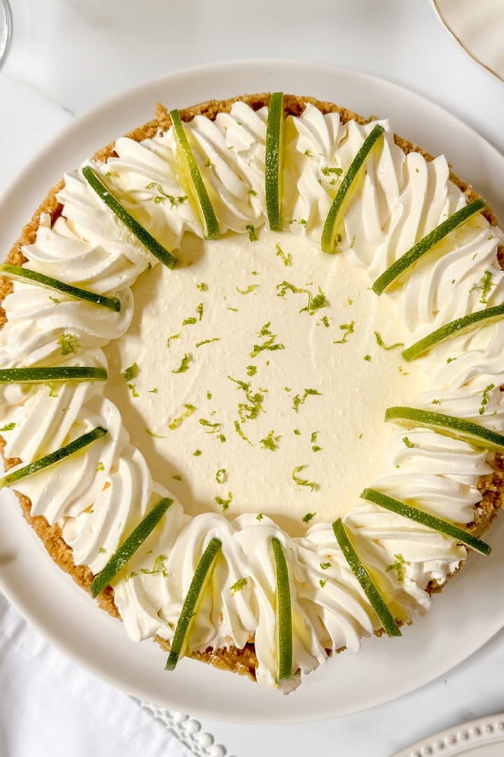 A ricotta cheesecake inspired key lime pie topped with whipped cream, lime wedges, and lime zest sits on a white plate on a marble surface.