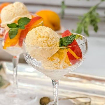 Two scoops of peach ice cream in a glass dessert cup, garnished with peach slices and a sprig of mint.