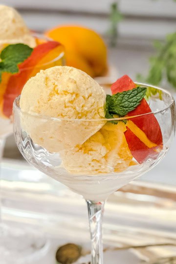 Two scoops of peach ice cream in a glass dessert cup, garnished with peach slices and a sprig of mint.