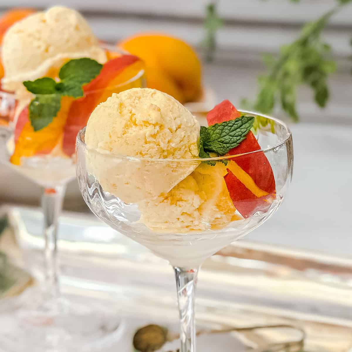 Two scoops of peach ice cream in a glass dessert cup, garnished with peach slices and a sprig of mint.