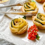 A puff pastry zucchini tartlet shaped like a rose with zucchini and yellow squash, served on a white plate with metal tongs and a small orange flower as garnish.