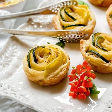 A puff pastry zucchini tartlet shaped like a rose with zucchini and yellow squash, served on a white plate with metal tongs and a small orange flower as garnish.