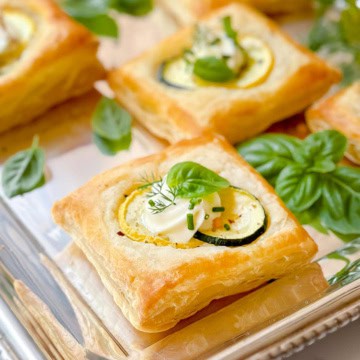 Square puff pastry savory tart topped with zucchini slices, cream, and fresh herbs, served on a silver tray alongside sprigs of basil.
