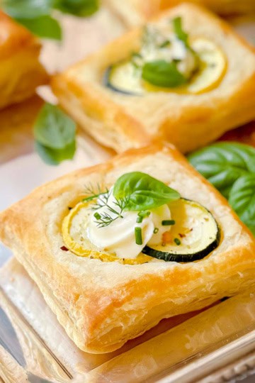 Square puff pastry savory tart topped with zucchini slices, cream, and fresh herbs, served on a silver tray alongside sprigs of basil.