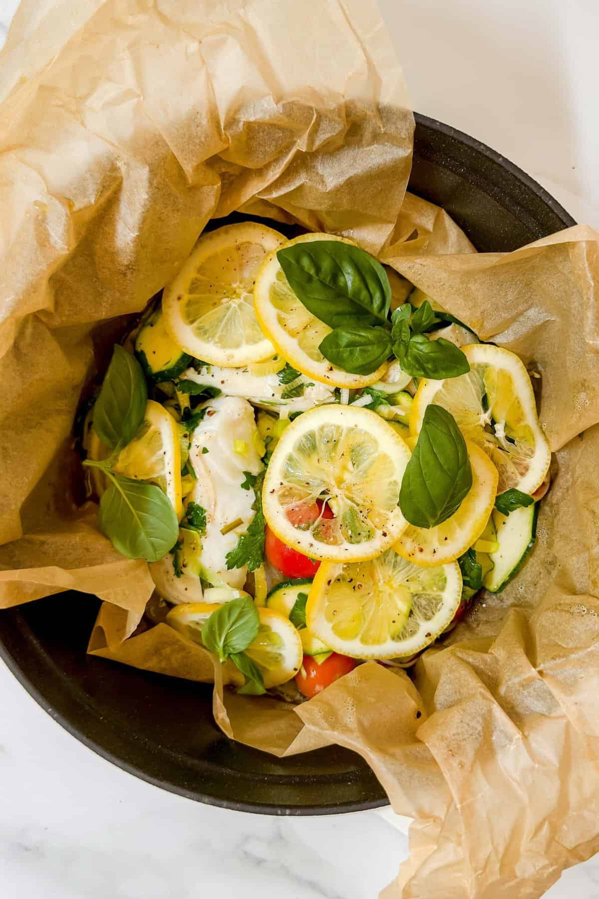 Chicken fillets topped with lemon slices, fresh basil leaves, and herbs, reminiscent of the delicate flavors found in steamed fish, placed on parchment paper in a baking dish.