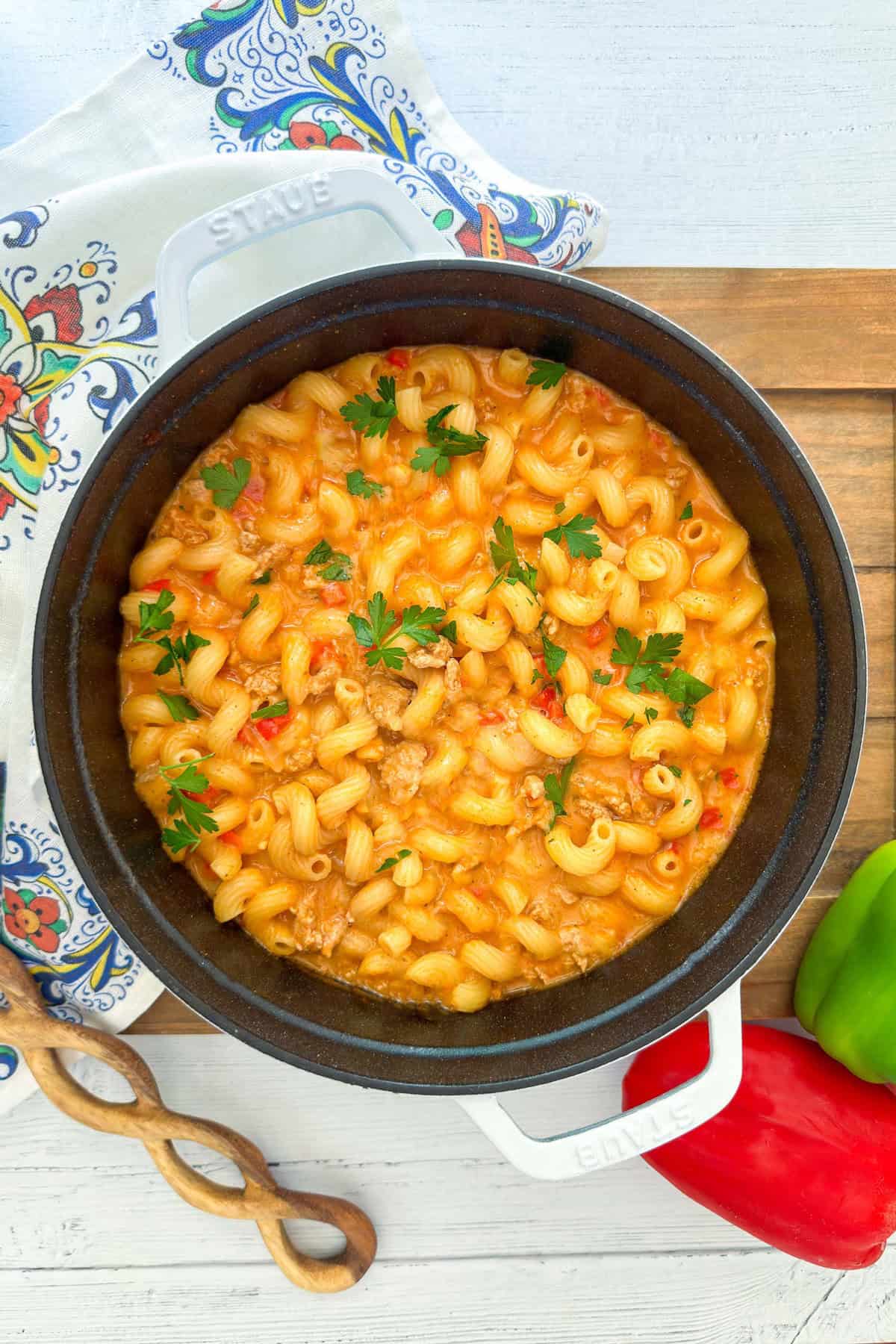 A pot of taco pasta with ground meat, tomato sauce, and chopped parsley on top sits on a wooden board beside green and red bell peppers.