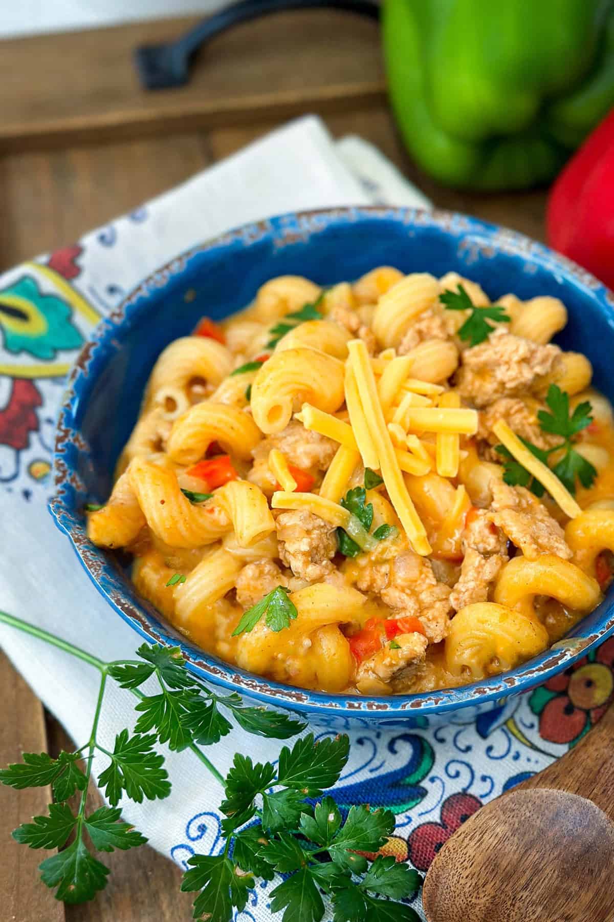 A blue bowl filled with cheesy taco pasta, ground meat, and parsley sits on a patterned napkin, with green bell pepper and parsley garnish nearby.