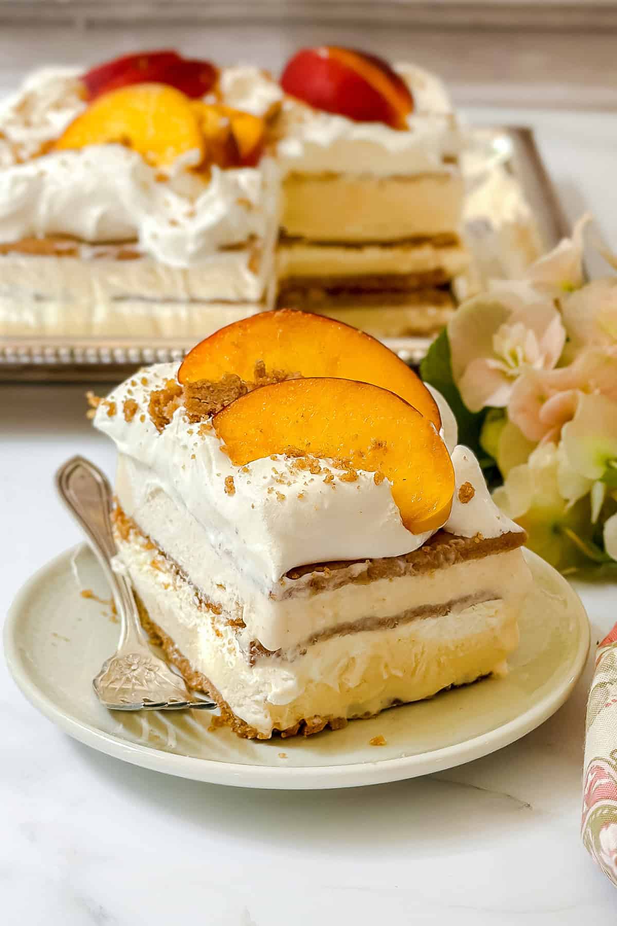 A slice of easy icebox cake layered to perfection is topped with whipped cream and fresh peach slices on a plate, with more cake and flowers in the background.
