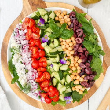 A large round platter with rows of chopped tomatoes, cucumbers, red onions, olives, chickpeas, feta cheese, and spinach&mdash;this stunning display celebrates the vibrant flavors of Mediterranean salads, garnished with fresh herbs and edible flowers.