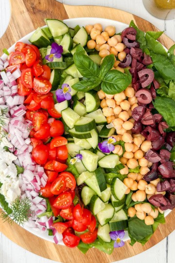A large round platter with rows of chopped tomatoes, cucumbers, red onions, olives, chickpeas, feta cheese, and spinach—this stunning display celebrates the vibrant flavors of Mediterranean salads, garnished with fresh herbs and edible flowers.